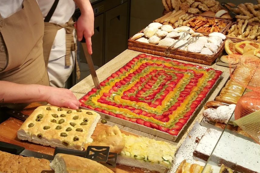 Le mani della fornaia tagliano una crostata di frutta, accanto ai cesti di paste e alla focaccia con le olive
